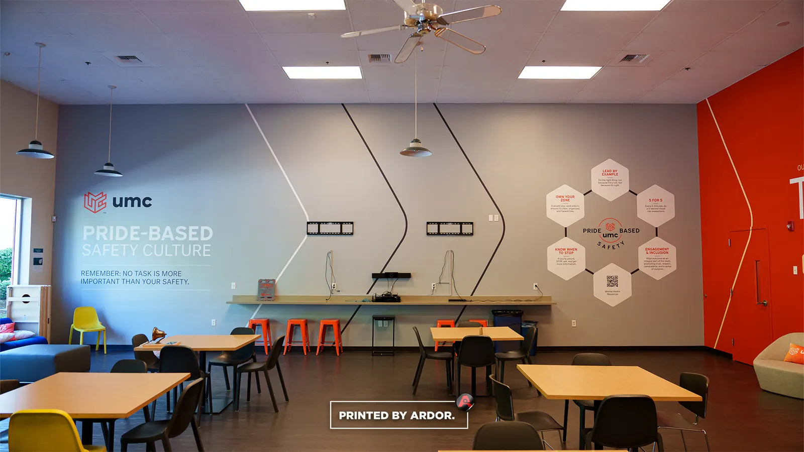 Employee break room with grey and orange wall wrap graphics about UMC pride-based safety culture above tables and chairs