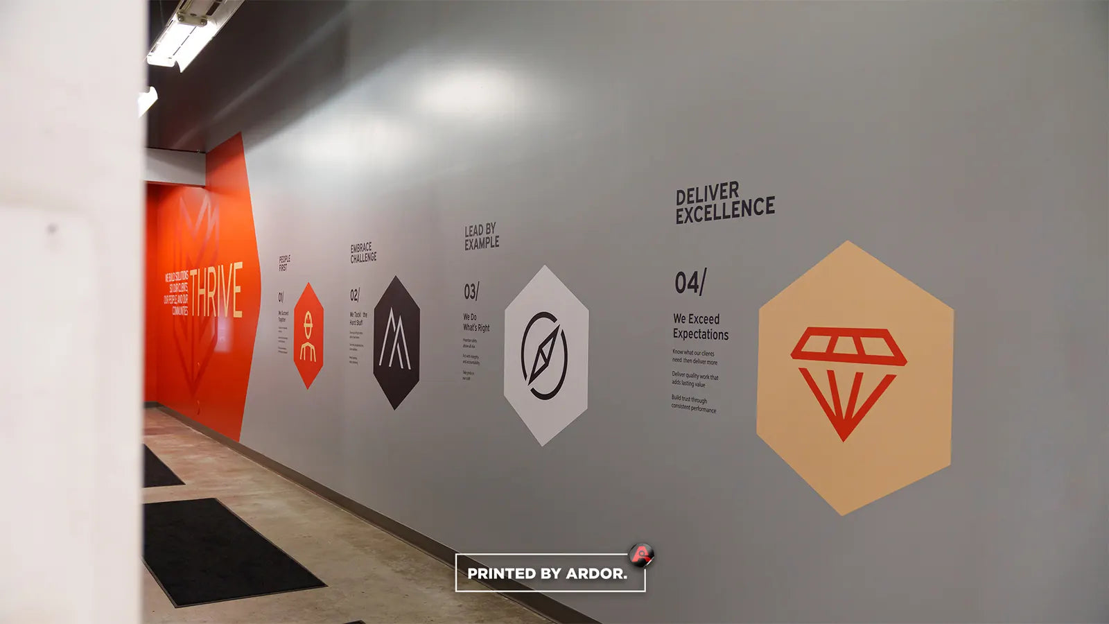 Office hallway wall wrap with orange and grey panels and hexagon icons listing core values such as Deliver Excellence