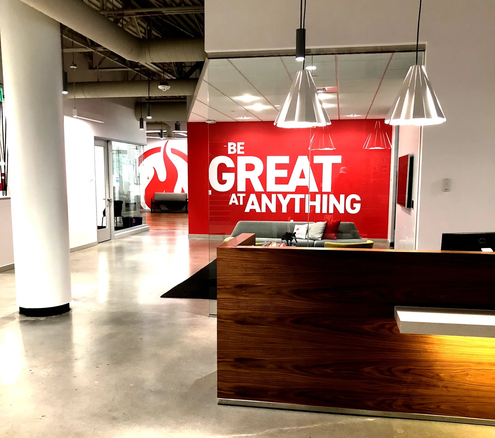 Red office lobby wall wrap with large white "Be Great at Anything" lettering behind a walnut reception desk with pendant lights