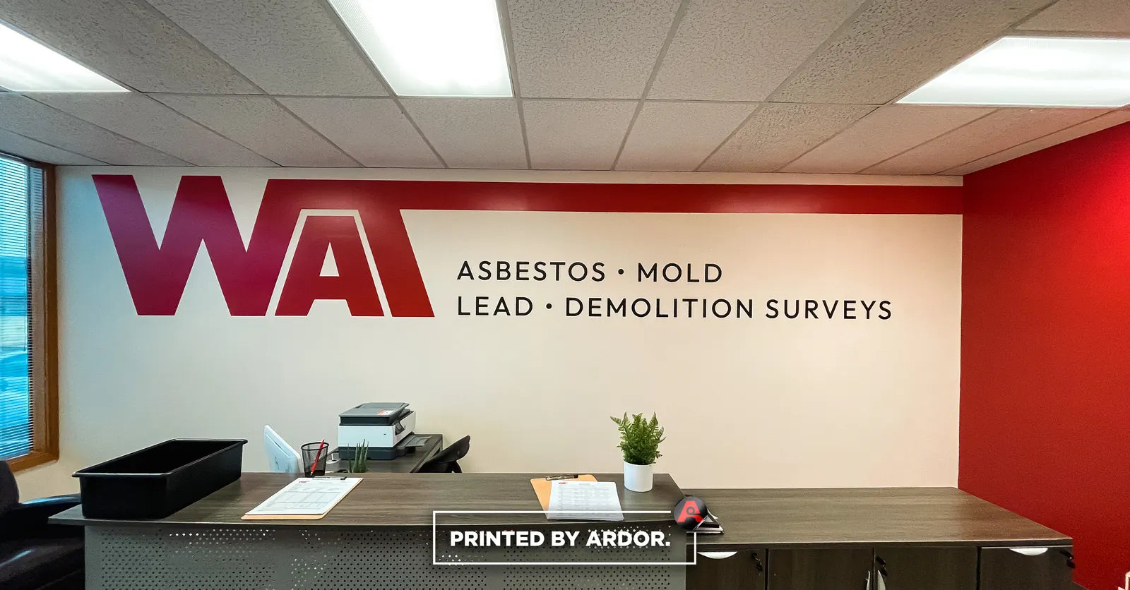 Reception desk in front of a white wall with a large maroon WAT logo and the words Asbestos, Mold and Demolition Surveys