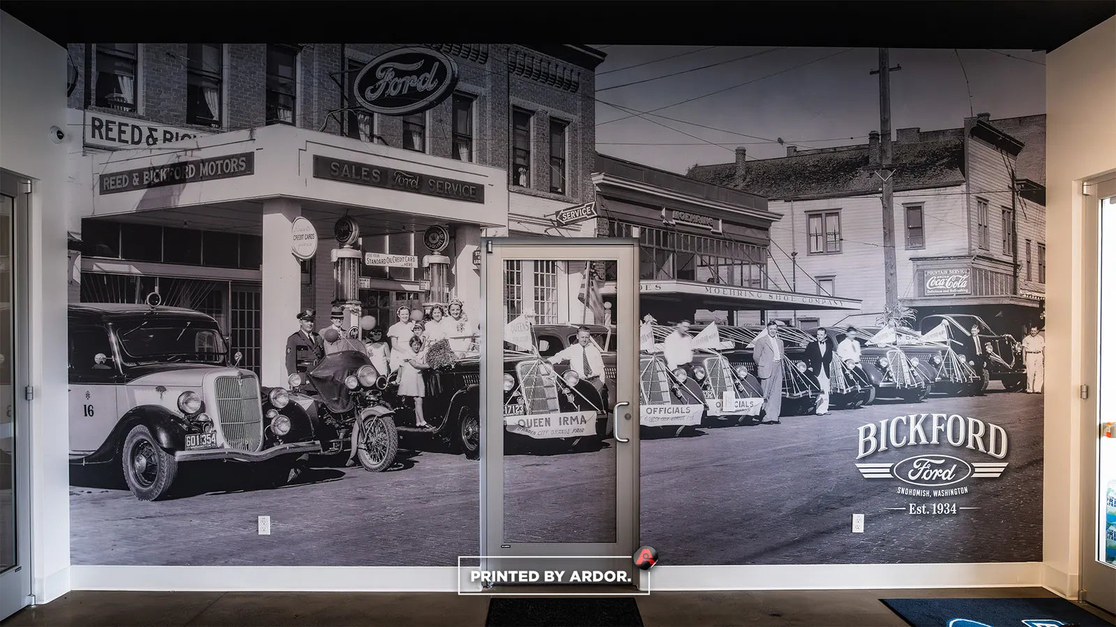 Black and white historic street photo mural of vintage Ford cars on a dealership wall with the Bickford Ford logo and a glass door