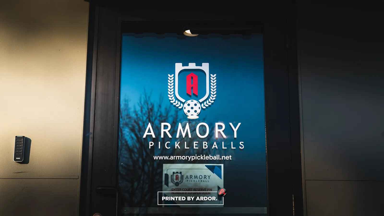 Blue wall graphic with the Armory Pickleballs crest logo and website printed beside a facility entrance door