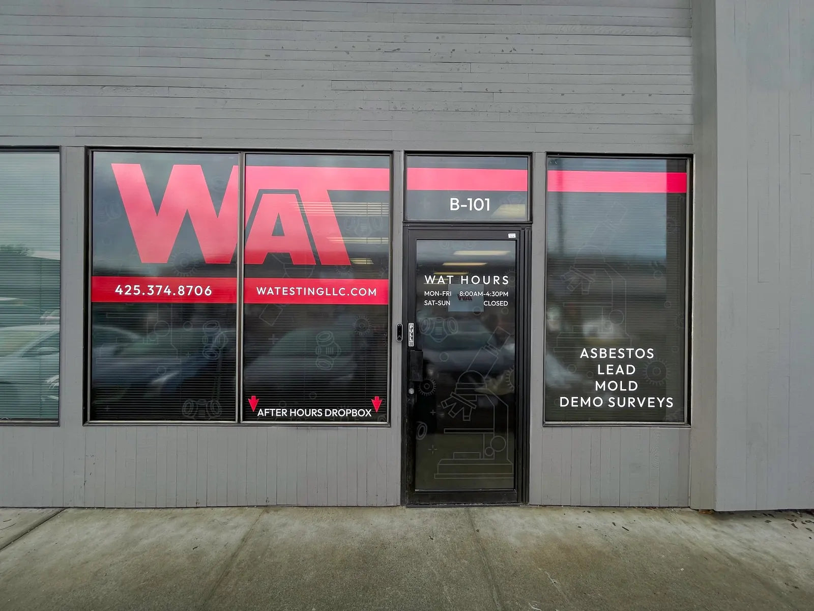 Storefront with perforated window film showing a large red WAT logo and phone number, plus business hours on the door
