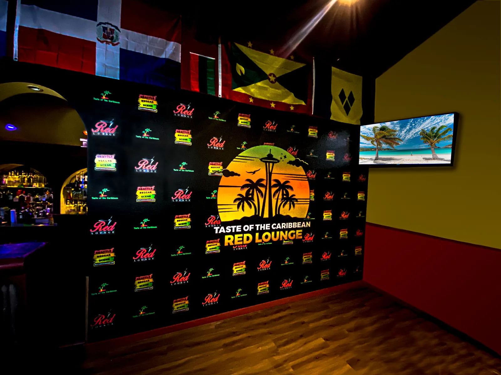 Black step-and-repeat style wall wrap with colorful Red Lounge logos and a palm tree and Space Needle sunset emblem, with flags and a TV above