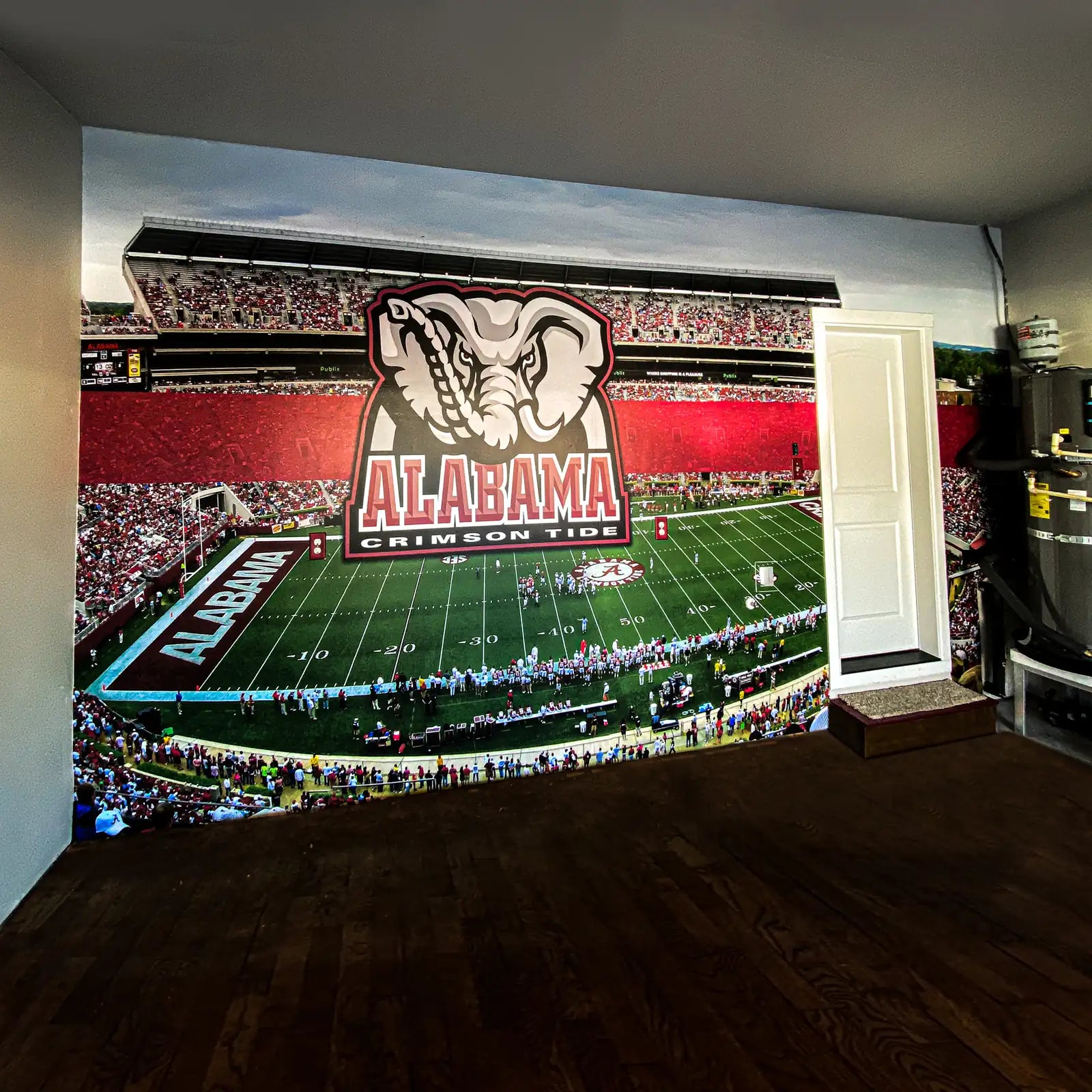 Full-wall photo mural of a packed football stadium with the Alabama Crimson Tide elephant logo, installed around a door in a finished room with a hardwood floor